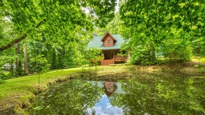 Une cabane entourée d’une végétation luxuriante, et un étang dans lequel se reflète la structure.