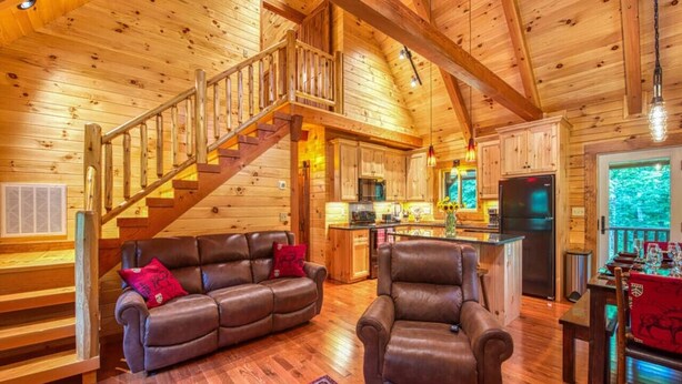 L ’intérieur d’une cabane en rondins comprend un escalier en bois, un espace de vie avec un canapé et un fauteuil, une cuisine et un coin repas avec une table et des chaises.