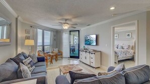 A living room with a sofa, chairs, a coffee table, and a TV. There is a bed in the background and a door leading to another room. - 308 Summerhouse by Avantstay Oceanview Condo w/ Community Pool Access (Isle of Palms)