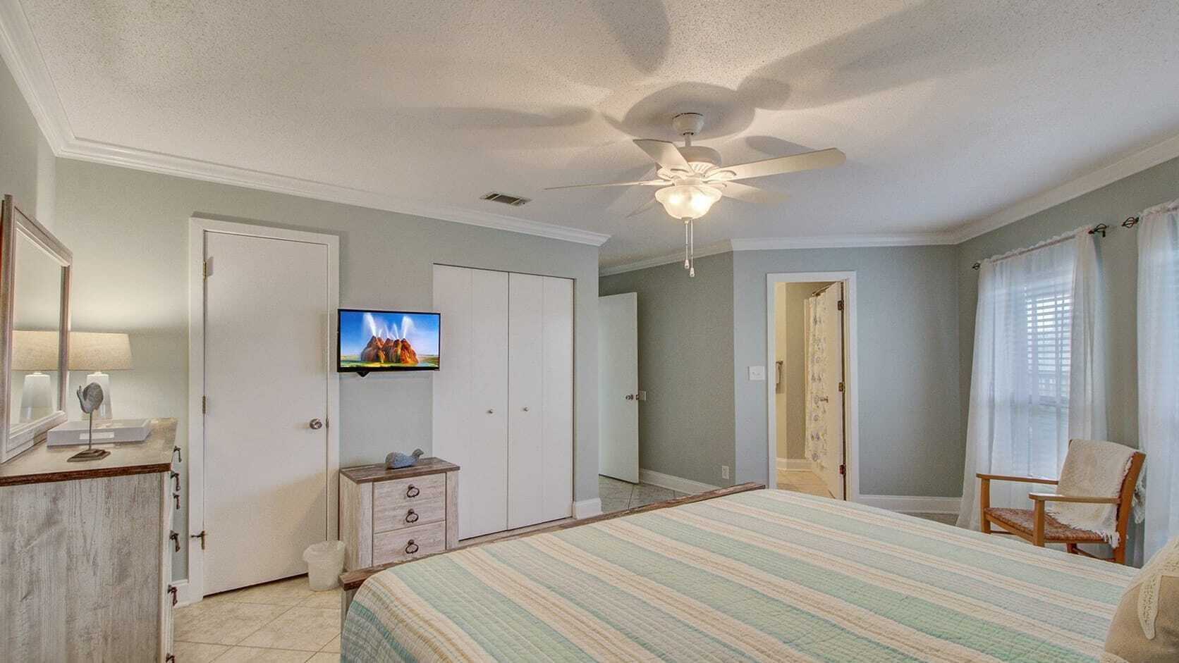 Un dormitorio con una cama, televisión, ventilador de techo y una silla.