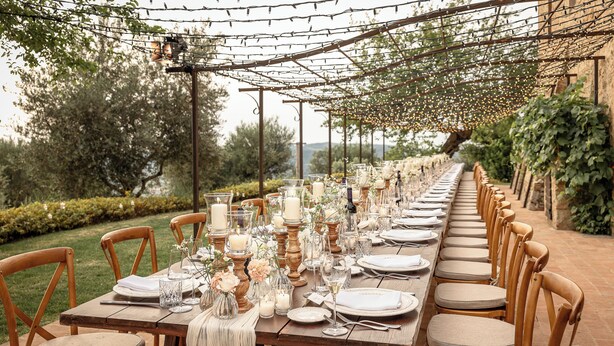 Une longue table en bois, dressée pour un événement formel en plein air, entourée de chaises en bois, et surmontée d’une pergola.