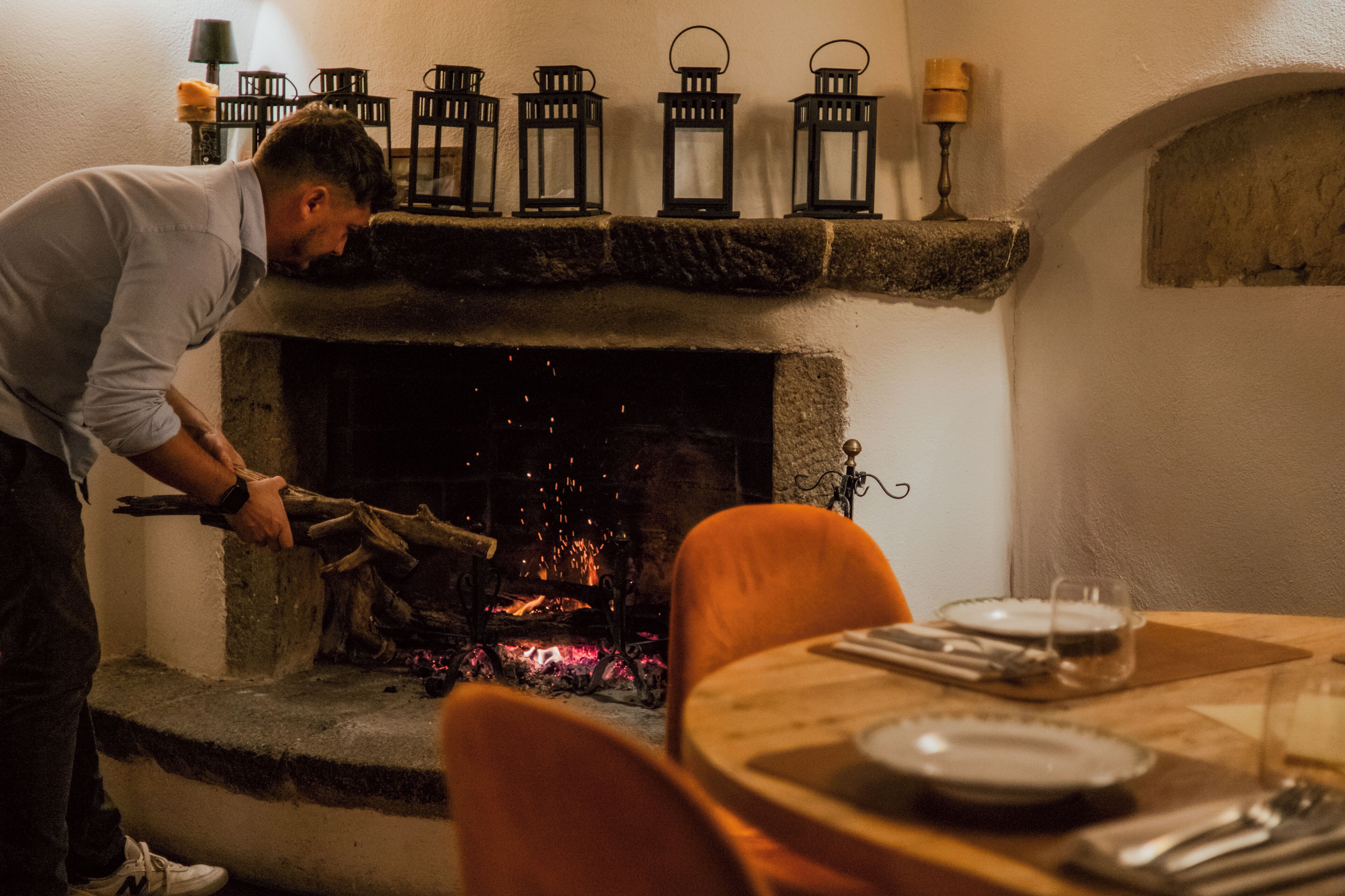 Une personne s’occupe d’un foyer avec des bûches, une table est dressée avec des assiettes et des couverts, et une chaise se trouve au premier plan.