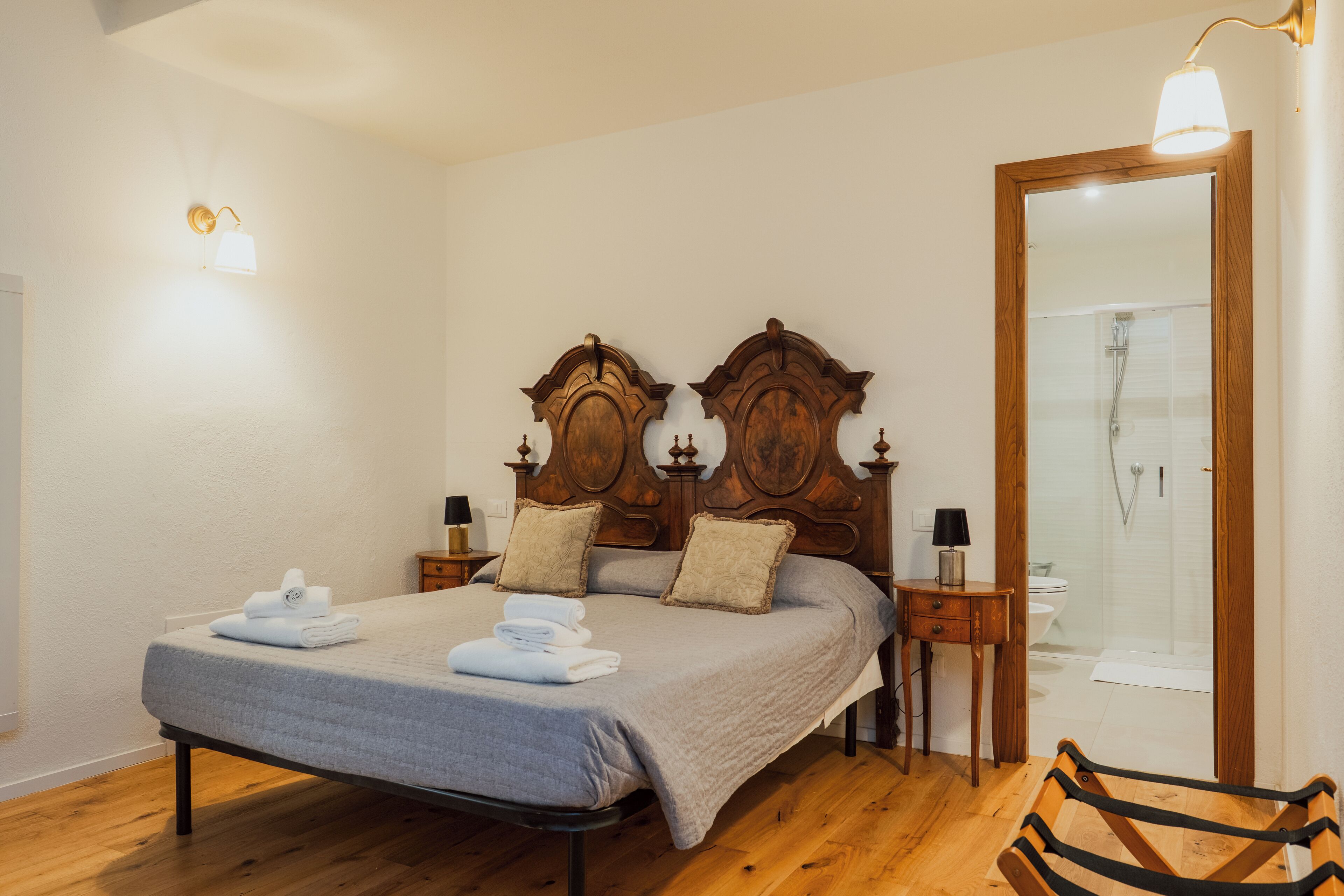 Una cama bien hecha con una colcha gris y dos almohadas, una mesita de noche de madera con una lámpara, y una silla de madera.