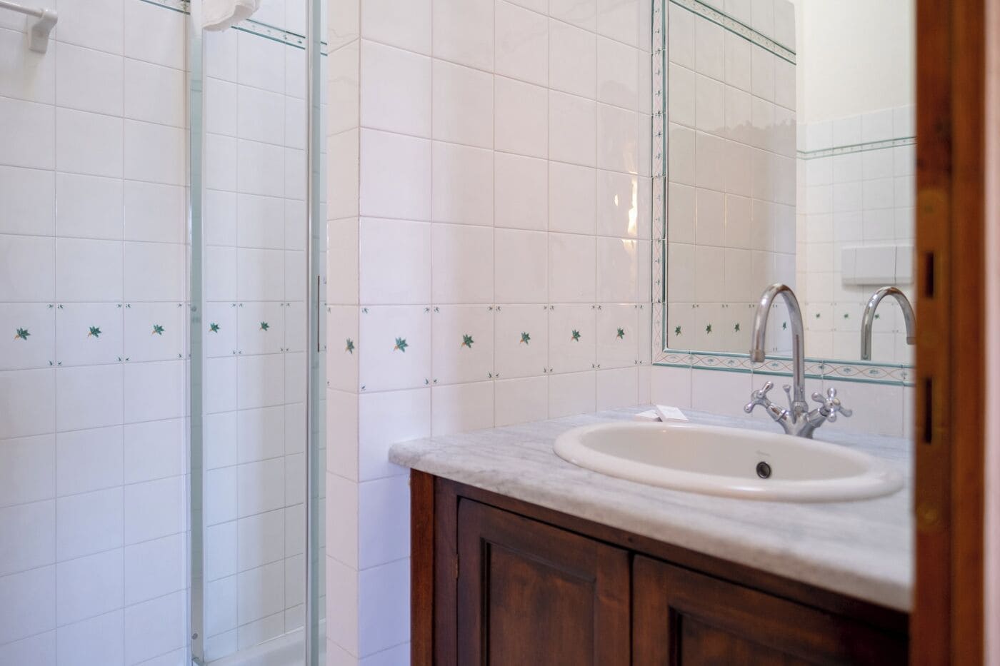 A bathroom with white tiled walls, a glass shower enclosure, a marble sink, and wooden cabinets.