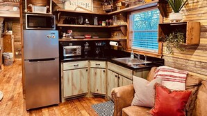 A compact kitchen with a built-in microwave, toaster, and sink. A 'Welcome' sign hangs above the counter, and there are wooden shelves with various kitchen items. - Cozy tiny cabin, kitchenette, private pond for fishing, near lake (Ringold)