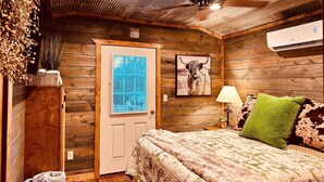 A cozy bedroom with a wooden ceiling, a ceiling fan, a bed with a floral quilt, a window with blinds, a framed picture of a yak, and a wall-mounted air conditioner. - Cozy tiny cabin, kitchenette, private pond for fishing, near lake (Ringold)