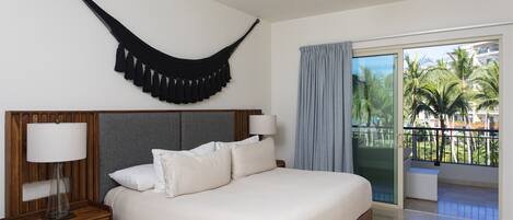 Habitación de hotel con una cama grande, una mesita de noche con lámpara, ventilador de techo y vista a palmeras a través de las puertas del balcón.