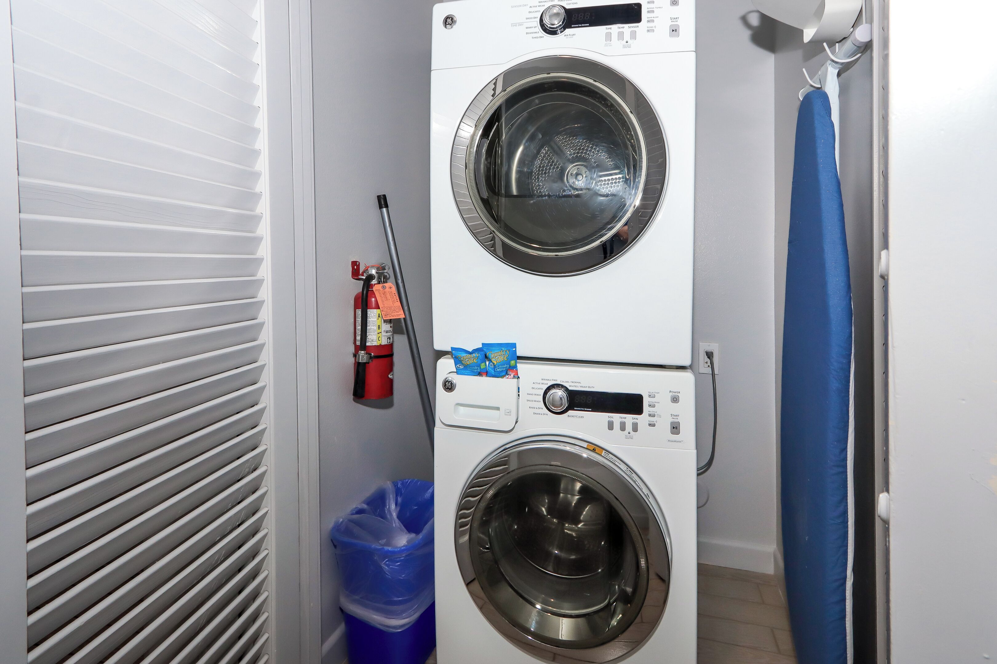 A front-loading washing machine with a control panel, a blue trash bin, and a fire extinguisher mounted on the wall.