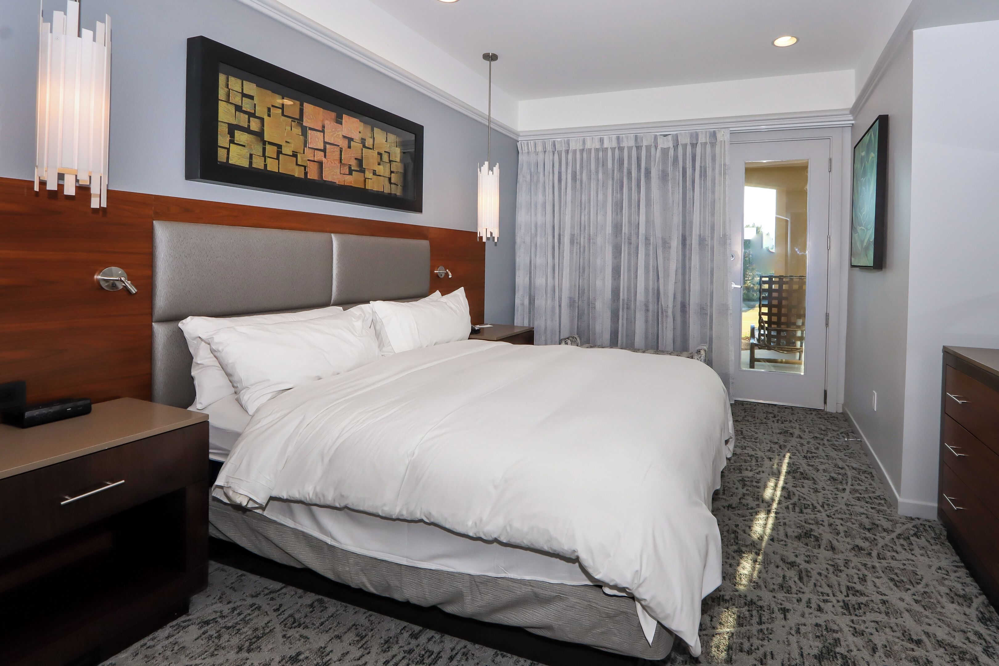 Quarto de hotel com uma cama grande, cabeceira de madeira, mesa de cabeceira, luminária na parede e vista para o exterior através de uma porta.