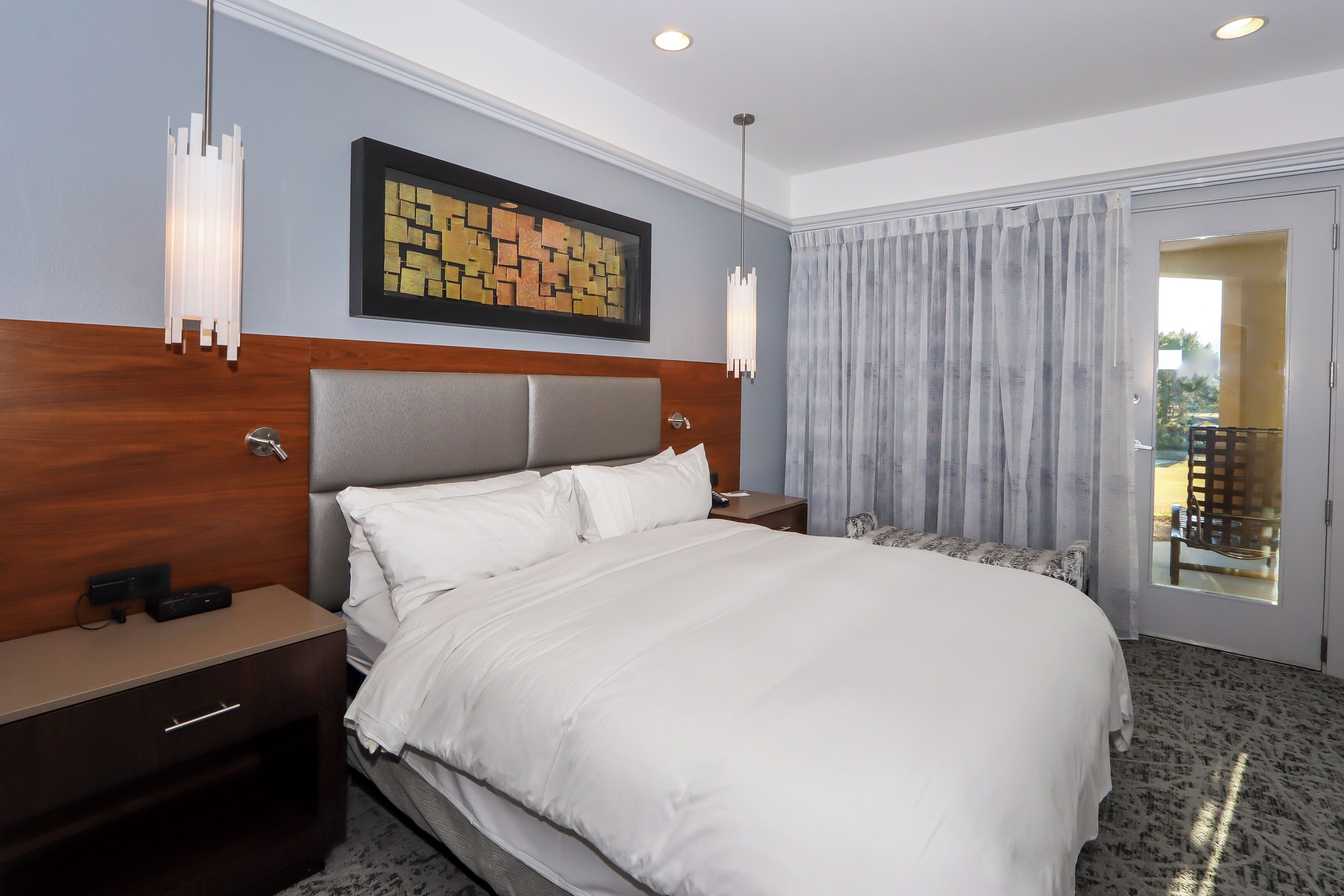 A hotel room with a large bed, a wooden headboard, a nightstand, a wall-mounted artwork, and a view through a door.
