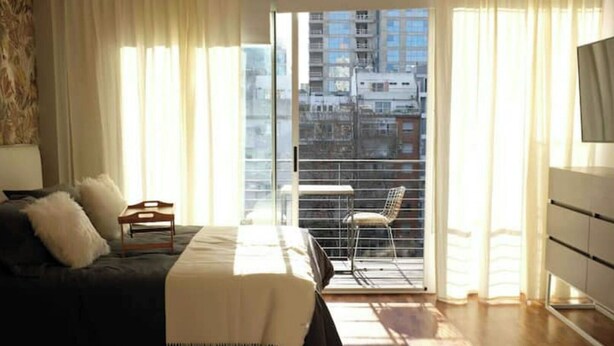 A bedroom with a bed, a chair, a TV, and a balcony with a city view. - Cozy Studio 7 Palermo Soho/ Balcony/Pool/Sum/AA. (Buenos Aires)