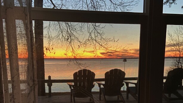 Vista desde una ventana hacia un atardecer sobre el agua, con dos sillas Adirondack en una terraza de madera.