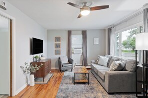 A modern living room with a flat-screen TV, a wooden cabinet, a grey sofa, a coffee table, and a ceiling fan. - Cozy Carriage Haus Close To Everything - Unit A (Cheyenne)