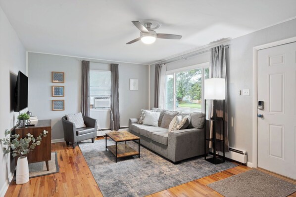 A modern living room with a grey sofa, a coffee table, a TV, and a ceiling fan. - Cozy Carriage Haus Close To Everything - Unit A (Cheyenne)