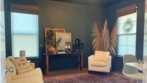 Ein Wohnzimmer mit einem gemusterten Teppich, einem weißen Sofa, einem weißen Sessel, einem kleinen Tisch mit Dekoration und hohen Pflanzen.