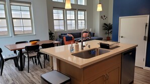 Una cocina moderna con una isla central, un comedor con mesa y sillas, y una sala de estar con sofá y planta.