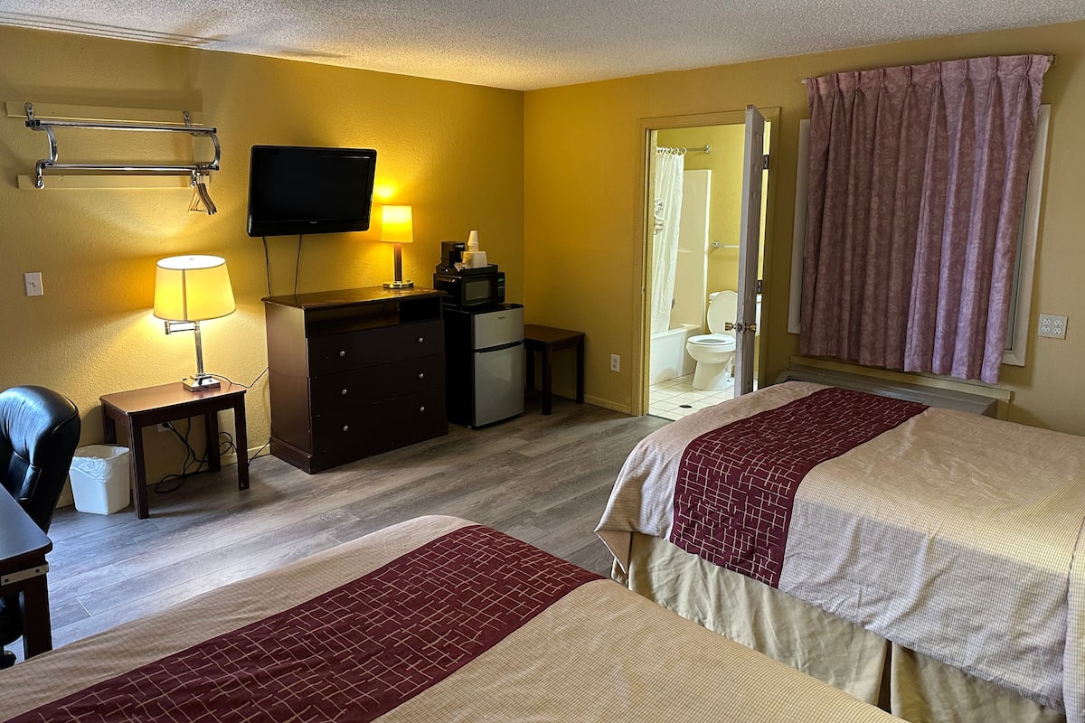 Habitación de hotel con dos camas, un escritorio, un televisor, un refrigerador y un baño visible a través de una puerta abierta.