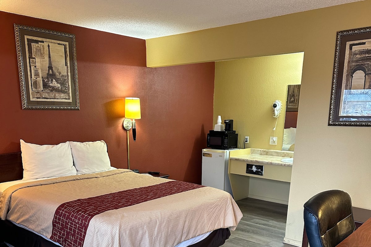 Habitación de hotel con cama, lámpara de noche, escritorio pequeño con silla, microondas y lavabo.