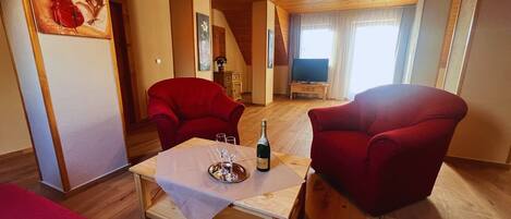 Una sala de estar con dos sillones rojos, una mesa de centro de madera con una botella y vasos, un televisor y un cuadro en la pared.