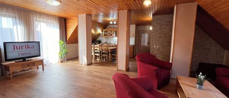 Un amplio salón con suelo de madera, sillones rojos, una mesa de centro de madera y un televisor mostrando 'Jurika Liptov'.