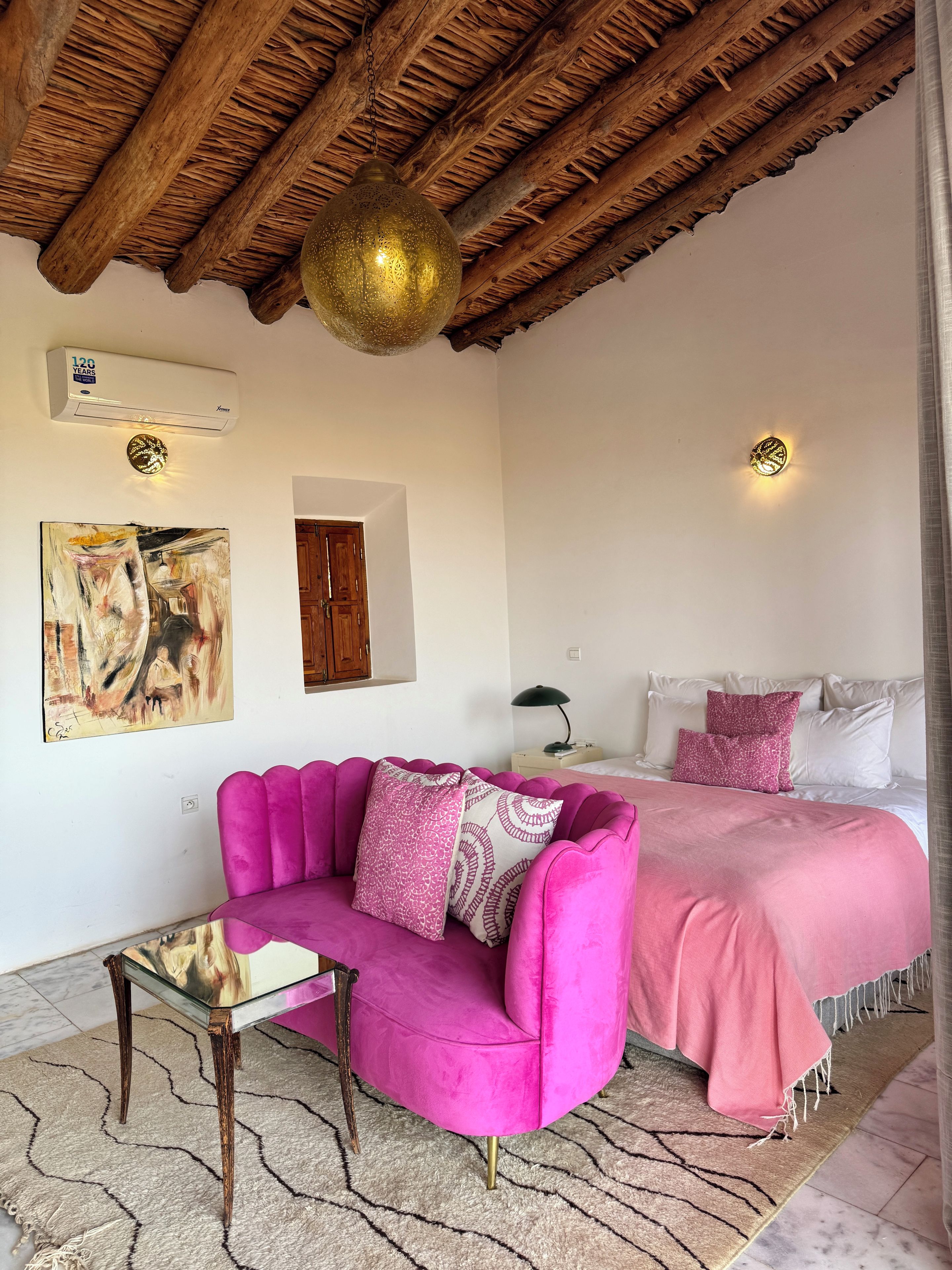 Habitació amb sostre de fusta, un sofà rosa, un llit amb llençols roses i un aire condicionat penjat a la paret.
