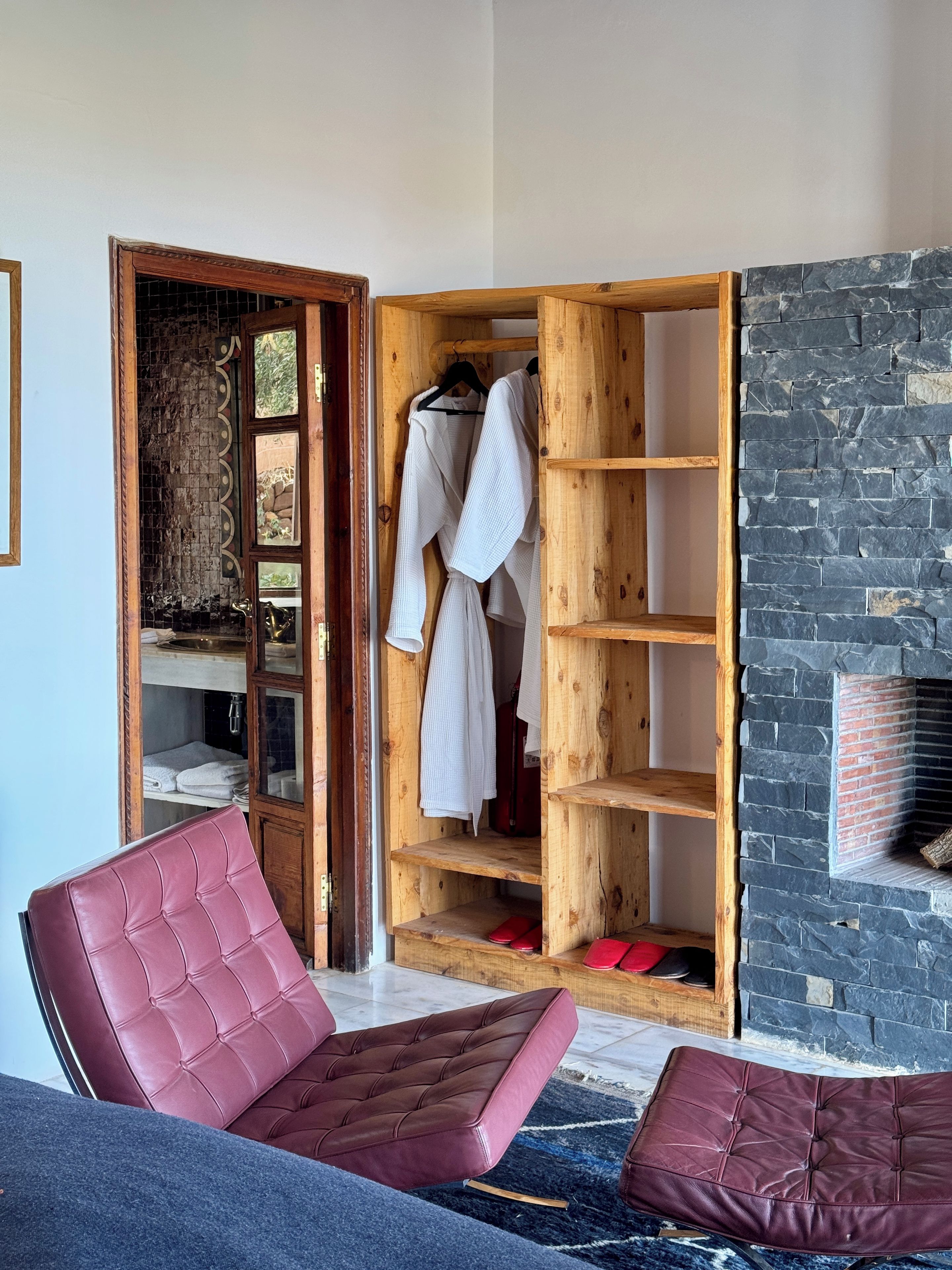 Una habitación con un armario de madera, una silla con cojín rojo y una pared de piedra.