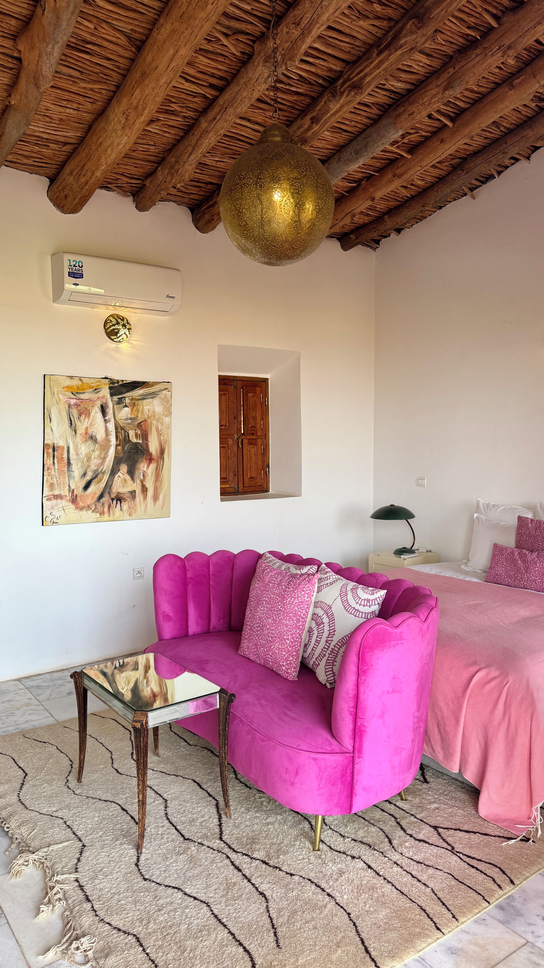 A room with a wooden ceiling, a pink sofa, a glass coffee table, a painting, and a bed with pink bedding.