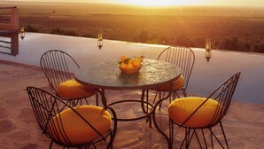 Mesa redonda com quatro cadeiras e uma tigela de fruta em um terraço com vista para uma paisagem ao pôr do sol.