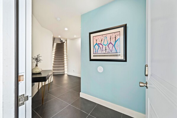 A hallway with a light blue wall, a framed abstract art piece, a staircase in the background, and a table with a vase.