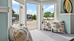 Quarto com vista para uma rua e carros estacionados, janela com vista para um prédio, e almofada estampada em um banco.
