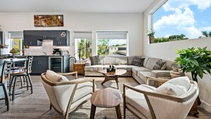 Uma sala de estar moderna com sofá, cadeiras, mesa de centro e uma cozinha ao fundo.