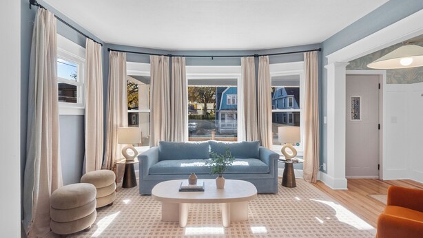 Ein Wohnzimmer mit einem blauen Sofa, einem runden Couchtisch und Blick auf ein blaues Haus durch ein großes Fenster.