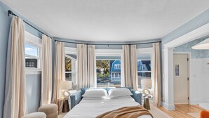 Ein Schlafzimmer mit einem großen Bett, zwei Nachttischen, einem Fenster mit Vorhängen und Blick ins Freie.