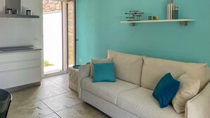 Een woonkamer met een beige bank, een blauwgroene wand en een keuken met een wit aanrecht.