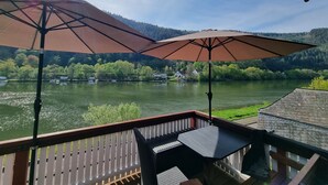 A deck with two umbrellas, a table, and a view of a lake with boats and houses in the background.