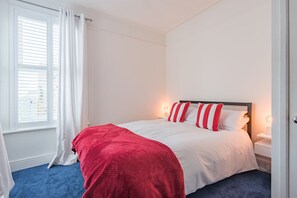 Un lit bien fait, avec une literie blanche et rouge, une moquette bleue et une fenêtre ornée de rideaux blancs.