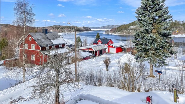 Un paysage rural avec une grange rouge et une maison, entourés d’arbres couverts de neige et d’une rivière gelée.