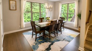 Comedor con mesa y sillas de madera, ventanas amplias con cortinas, candelabro y alfombra estampada.