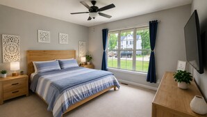 Un dormitorio con cama, mesitas de noche, escritorio, televisor, ventilador de techo y ventana con cortinas.