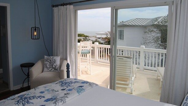 Une chambre à coucher avec un lit, une table de chevet, une chaise et une vue sur un balcon doté d’une balustrade, avec une maison en arrière-plan.