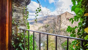 Varanda com parede de pedra, grade de metal e vista para paisagem montanhosa com um edifício ao longe.