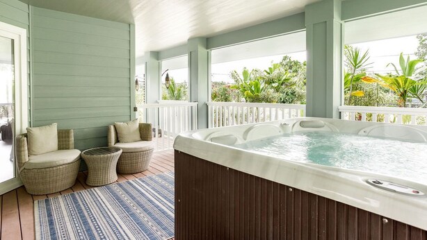 A covered outdoor area with wicker furniture, a hot tub, and a view of greenery. - NEW - Lauhala - Hawaiian Villa with private hot tub (Keaau)