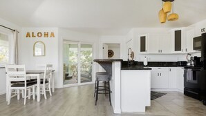 A modern kitchen with white cabinetry, a central island, and a dining area with white chairs. - NEW - Lauhala - Hawaiian Villa with private hot tub (Keaau)