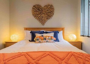 Cama bem arrumada com decoração em formato de coração na parede, duas luminárias de cabeceira e uma janela com persianas.