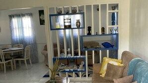 Una sala de estar con un sofá, una mesa de centro, una mesa de comedor con sillas y una estantería decorativa con diversos objetos.