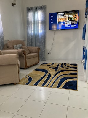 Un soggiorno con due poltrone beige, un tappeto a motivi, una televisione a parete che mostra i telegiornali e una finestra con tende leggere.
