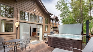 Une terrasse en bois avec un jacuzzi, des sièges d’extérieur et une vue sur une maison dotée de grandes fenêtres et d’un balcon.