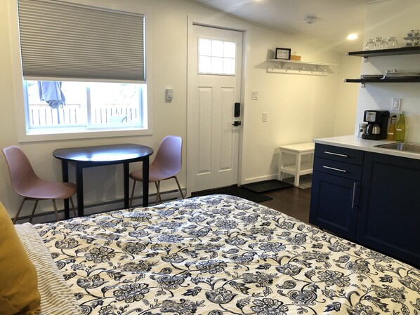 A small, well-lit room with a bed, two chairs, a table, a window with blinds, a door, a small white table, a coffee maker, and shelves with glassware.