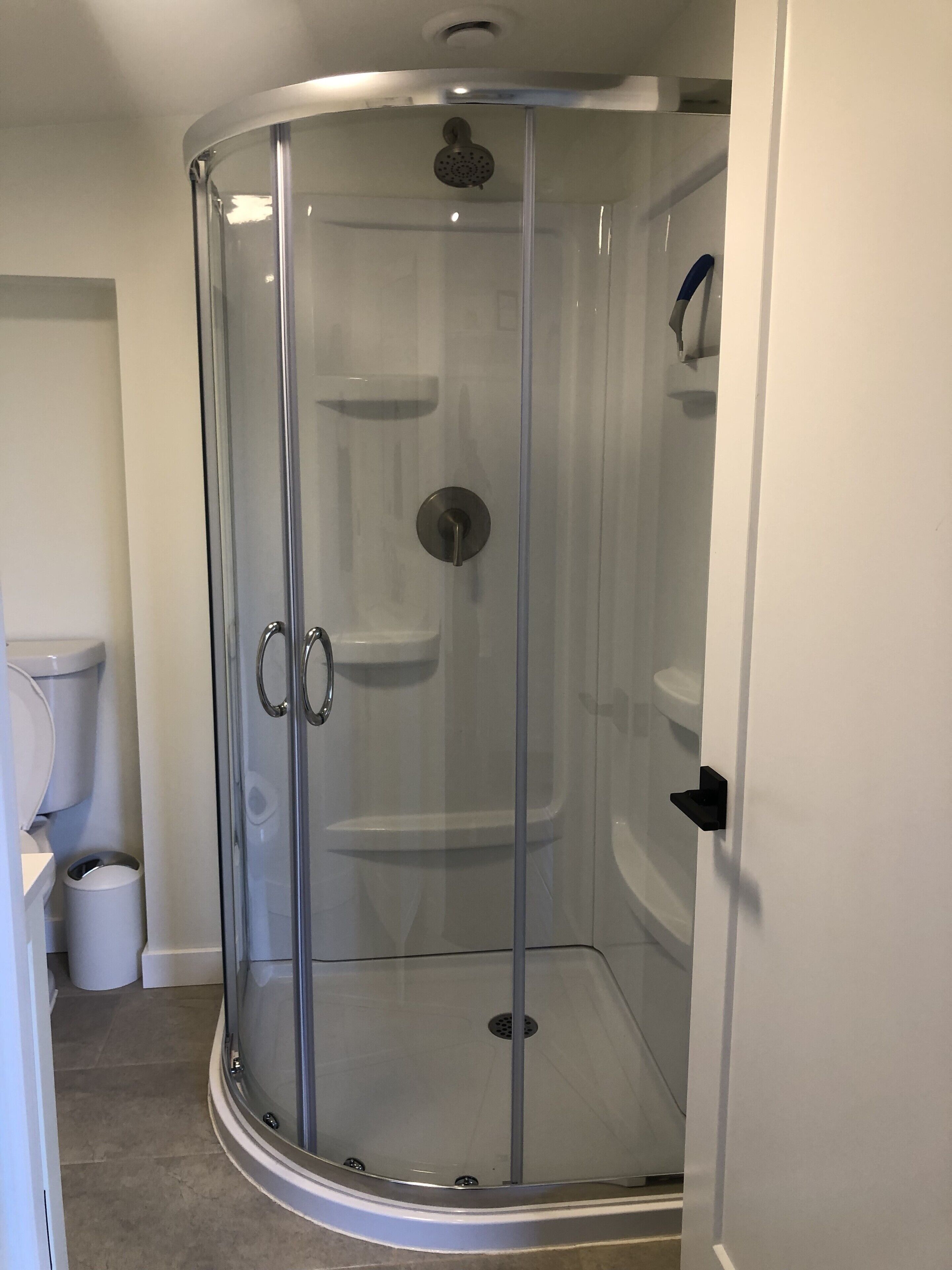 A glass-enclosed shower stall with a door, a toilet, and a trash can in a bathroom.
