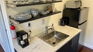 A kitchen with a marble countertop, stainless steel sink, and a coffee maker. There are shelves above the counter holding plates, bowls, and cups. A toaster and a refrigerator are also visible. - Bachelor Suite Bed & Breakfast in Northern Muskoka (Huntsville)